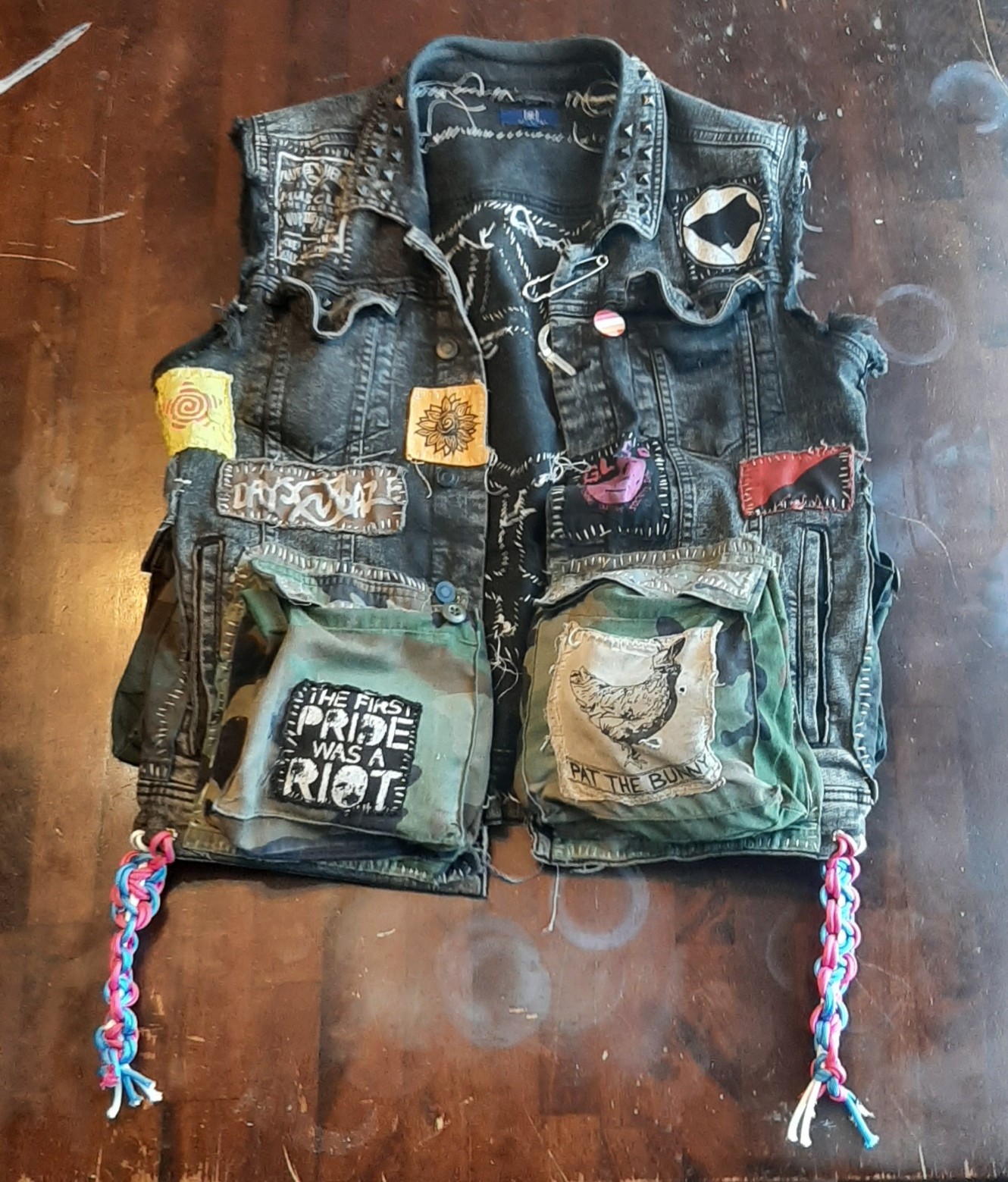 back of the Vest Of Holding, my black denim punk vest, covered in patches and pins and studs. the reflective tape tranarchy symbol glows brightly beneath the camera flash. paracord tassels in trans pride colors hang down the sides